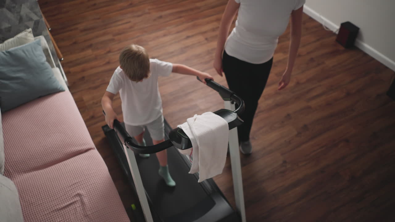 Overhead view shows fitness trainer directing young boy on treadmill workout as he looks down, focusing on steps, with pink couch, wooden floor, and home interior setting in background