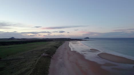 imágenes de drones volando lentamente hacia atrás sobre una larga playa de arena y dunas de arena durante una puesta de sol rosa mientras la marea golpea suavemente la orilla y la gente camina por la playa