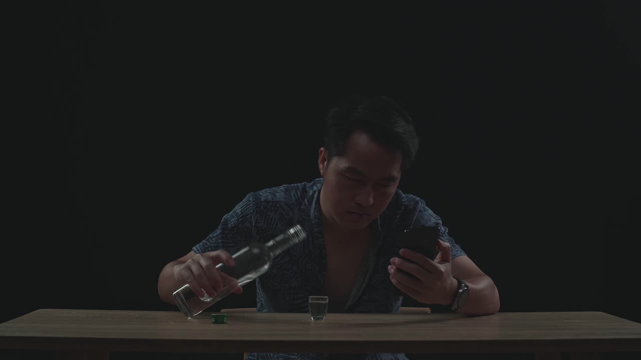 술에 취한, 우울한 아시아 남자가 검은 바탕의 스마트폰을 사용하는 동안 유리잔에서 보드카를 마시고 있습니다.