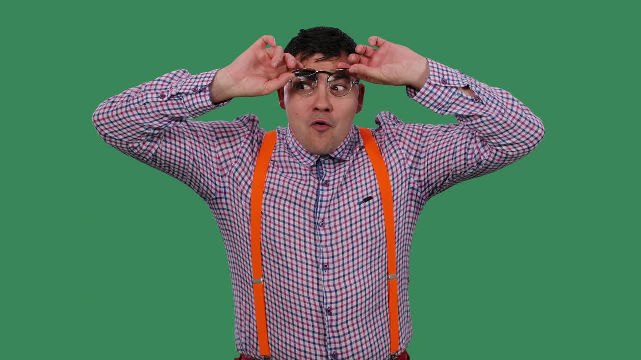 선글라스를 입은 남자가 얼굴에 환상적인 표정으로 색의 렌즈를 들어 올립니다. 초록색 스크린에 있는 스튜디오에서 셔츠를 입은 안경을 입은 남자의 초상화.