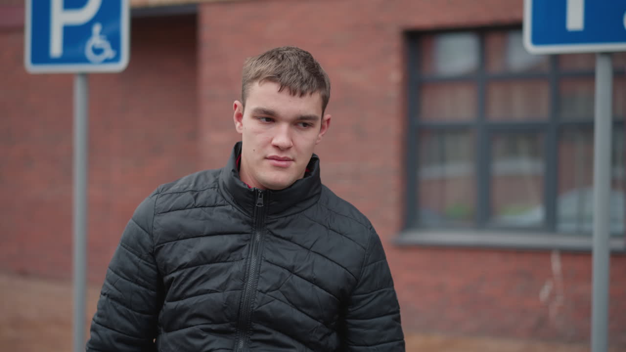 Front view of smiling young driver wearing black winter coat and blue jeans walking toward parked car on urban pavement with red brick buildings in background during cold city day