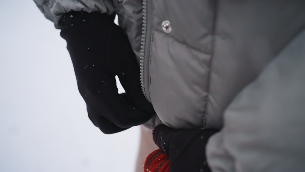 Hand in black glove pulling zipper on gray winter coat in snowy outdoor setting, showcasing cold weather attire and subtle hand motion, capturing moment of preparing for warmth in forest environment