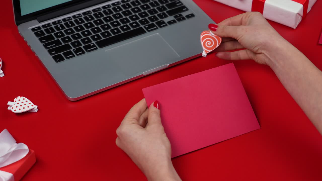 Woman's hands open envelope and take out valentine's love postcard with a green screen chroma key. Top view of a red table with a laptop, hearts and gift boxes. Valentine's Day. Close up. Slow motion