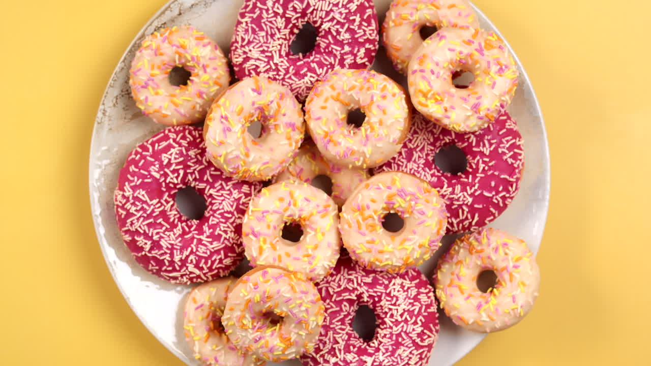 Closeup of Female Hand putting small Donut on Plate full of colorful doughnuts in different size, pink and white on light yellow background. Top view, filmed in Prores.