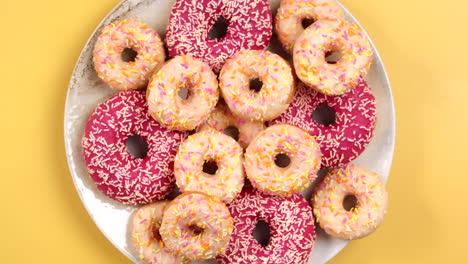 closeup of female hand putting small donut on plate full of colorful doughnuts in different size, pink and white on light yellow background. top view, filmed in prores.