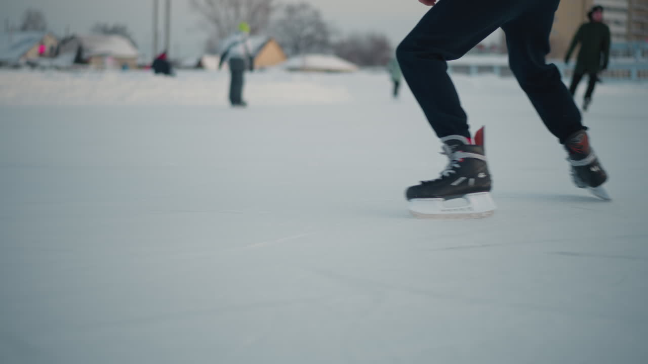 lower angle view of person skillfully skating on frozen lake with other blurred skater in background, ice surface smooth with blade marks, cold winter outdoor scene under soft light