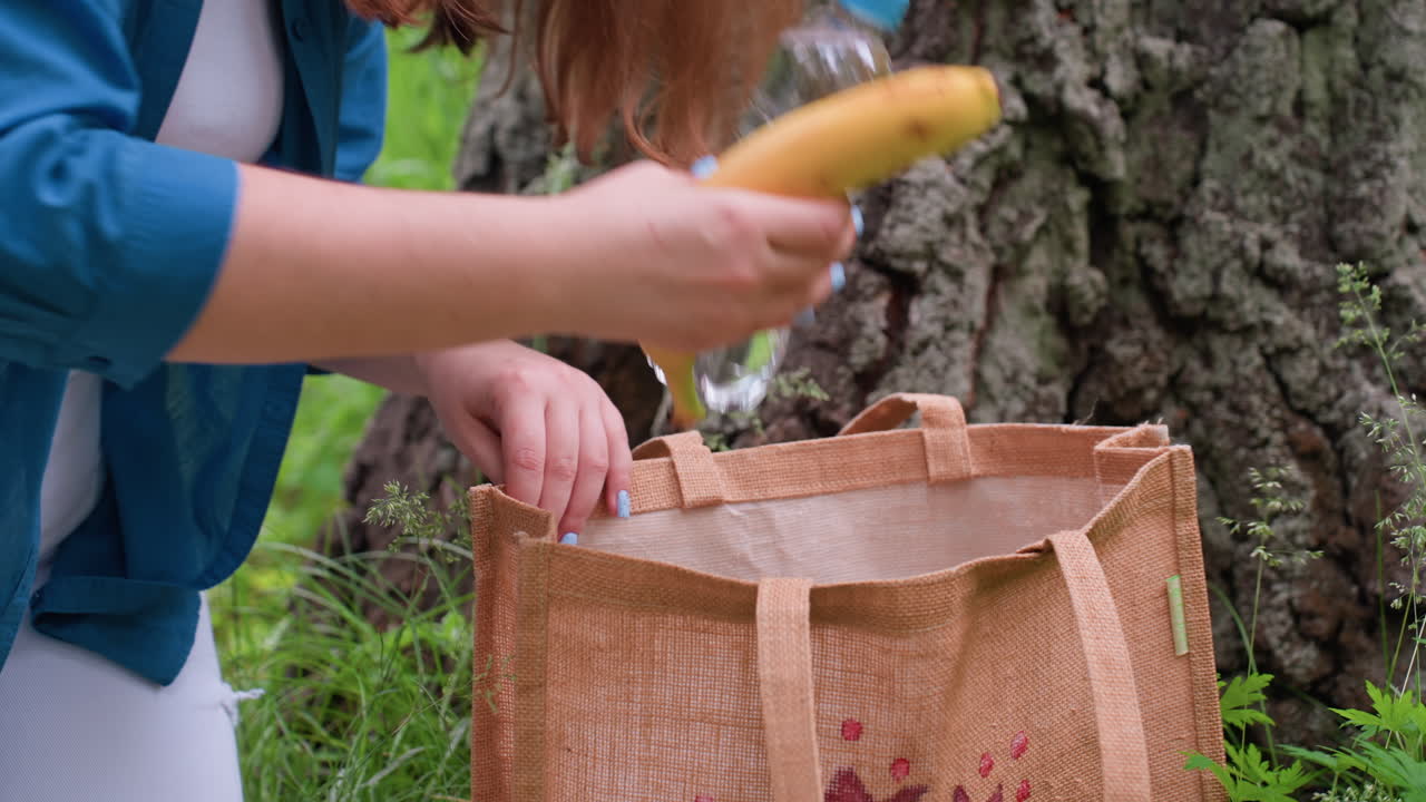 Close up of woman with golden hair returning banana and bottle water into brown fabric bag beside tree trunk, surrounded by green grass and sunlight