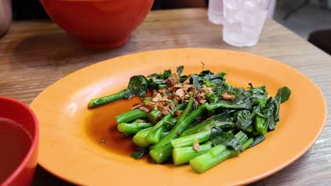 a hand uses red chopsticks to pick up chinese broccoli from an orange plate in a casual singaporean eatery, under bright, natural lighting