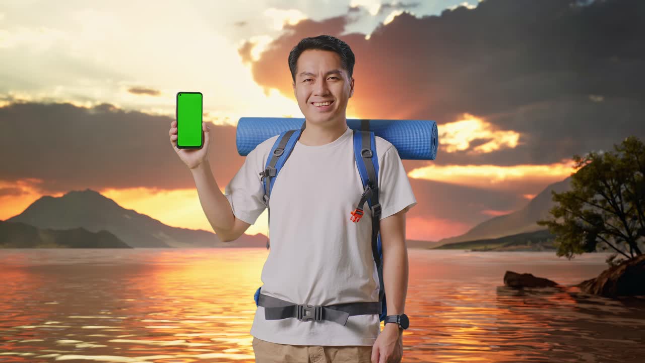 excursionista masculino asiático con mochila de montañismo sonriendo y mostrando una maqueta de teléfono inteligente de pantalla verde en un lago