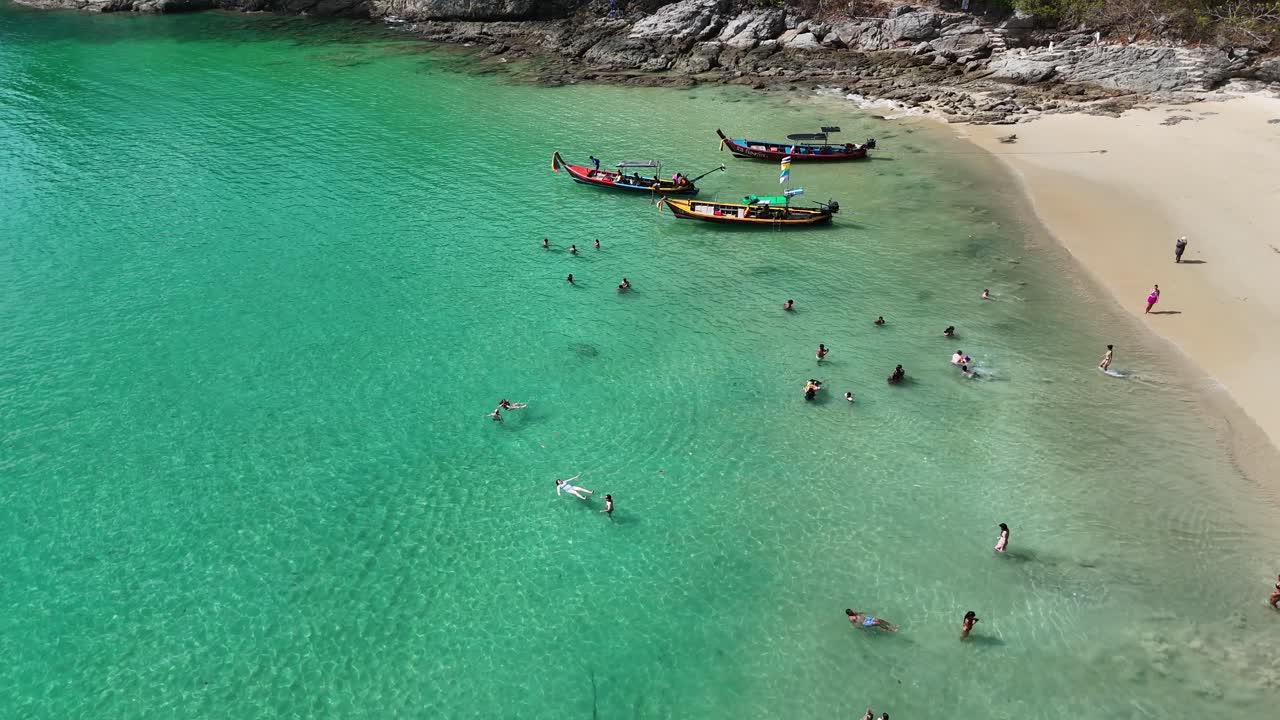 Drone footage captures people swimming near longtail boats on a sunny beach in Phuket, Thailand, showcasing vibrant turquoise waters
