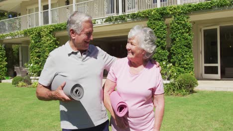 feliz pareja de ancianos caucásicos haciendo ejercicio abrazándose caminando llevando esteras de yoga en el jardín al sol