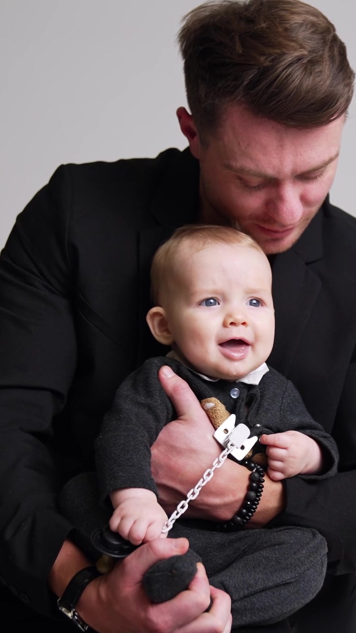 Uomo che abbraccia un bambino piccolo. Padre in giacca nera tiene in braccio il figlio neonato e lo bacia amorevolmente sulla testa. Riprese in studio. Sfondo bianco. Video verticale.