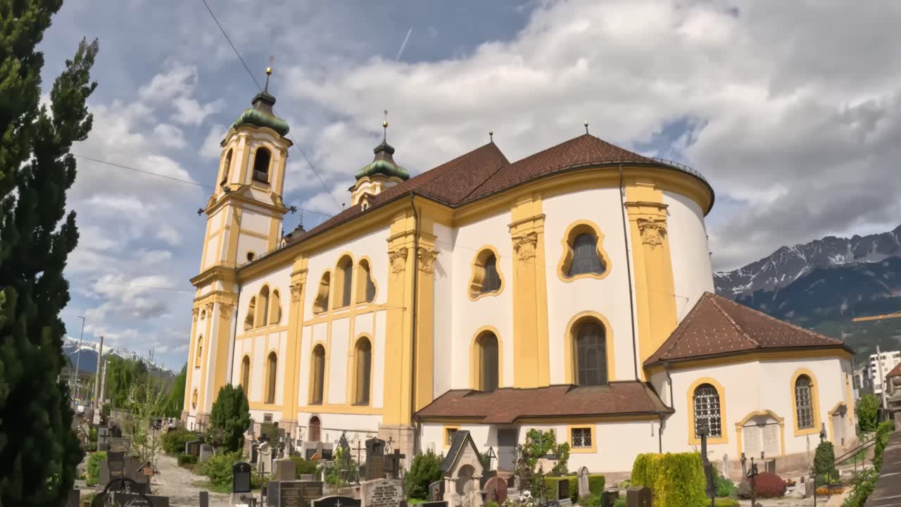 Wide-angle establishing shot of Pfarrkirche und Basilika Mariae Empfängnis in Innsbruck, Austria, showing its yellow baroque façade, nearby cemetery, and snow-capped Alps in the background