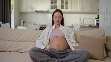 A-pregnant-brunette-woman-in-gray-pants-and-a-white-shirt-sits-on-the-sofa-and-meditates-at-home-while-expecting-a-baby