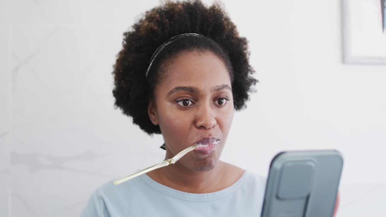 African american woman brushing teeth and using smartphone in bathroom, slow motion