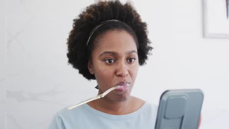 mujer afroamericana cepillándose los dientes y usando un teléfono inteligente en el baño, cámara lenta