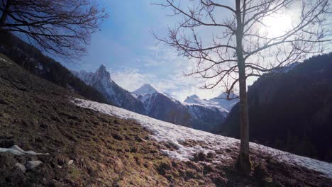 Snow-melting-on-a-hill,-beautiful-white-moutain-and-peak-in-the-background,-in-winter-on-a-blue-sky-bright-day,-with-sun-flares-and-beams