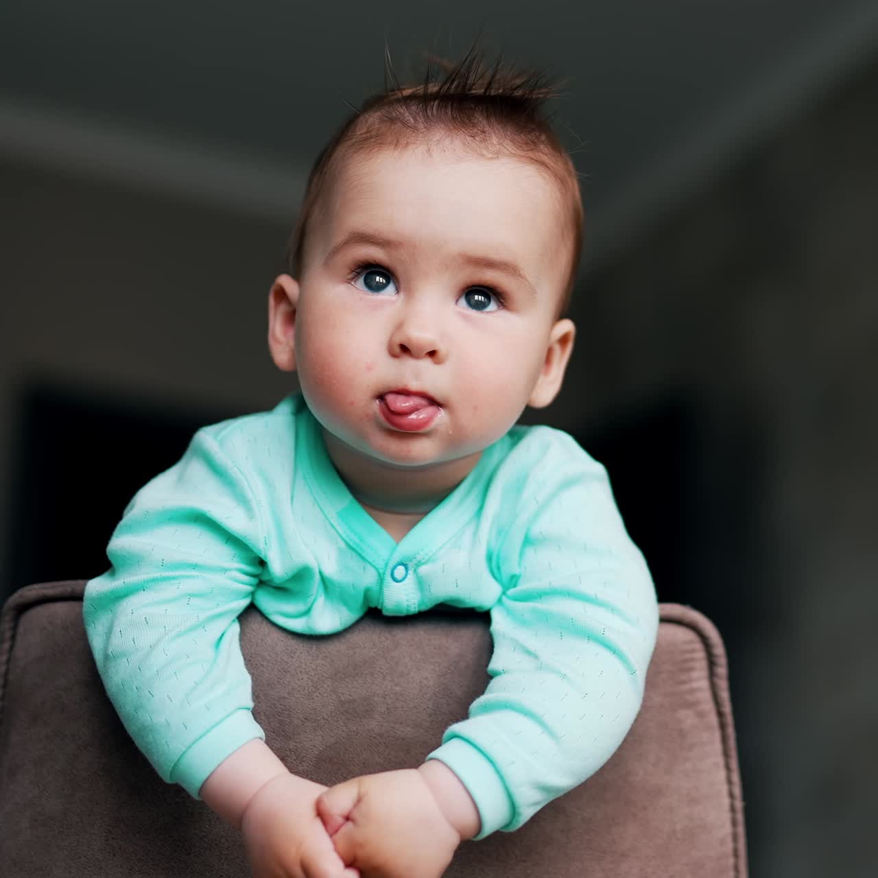 Morsom og nydelig unge med utstående hår står på stolen. Babyen ser fremover og rekker tunge på en leken måte. Lav vinkel.