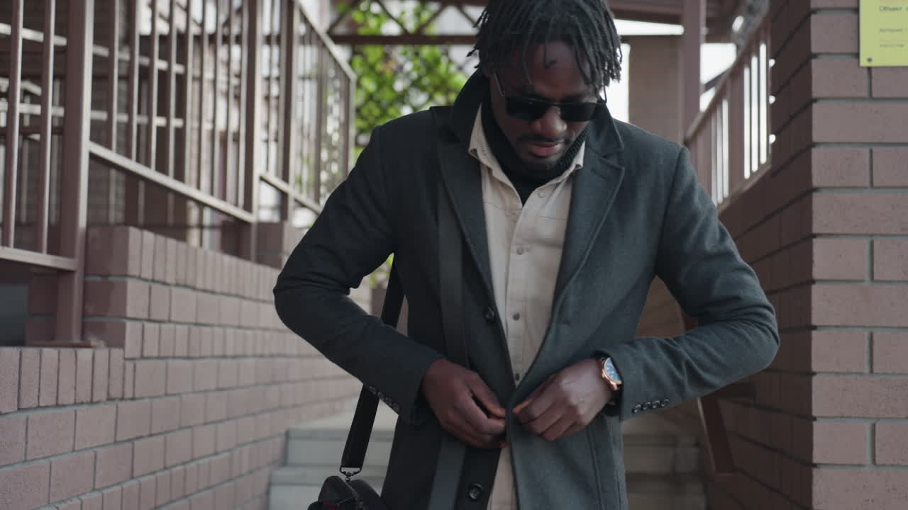 Un homme moderne portant des lunettes de soleil ajuste son manteau en descendant un escalier extérieur, avec des balustrades en brique et un treillis derrière lui, une montre-bracelet qui brille, une démarche assurée, une entrée de campus urbain aux couleurs discrètes et à l’ambiance moderne.