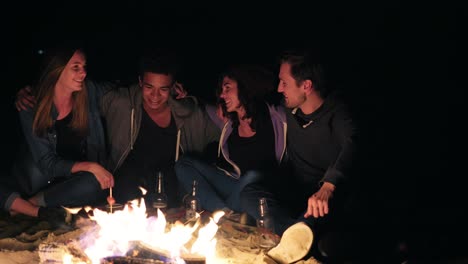 Diverse-group-of-people-sitting-together-by-the-fire-late-at-night-and-embracing-each-other,-cooking-sausages-and-drinking-beer.-Cheerful-friends-talking-and-having-fun-together