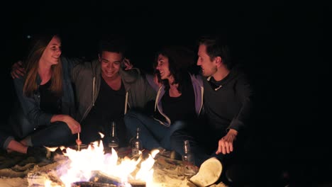 grupo diverso de personas sentadas juntas junto al fuego tarde en la noche y abrazándose, cocinando salchichas y bebiendo cerveza. amigos alegres hablando y divirtiéndose juntos