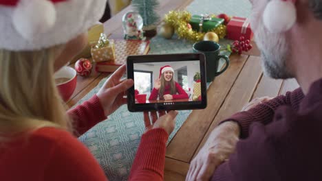 Lächelndes-Kaukasisches-Paar-Mit-Weihnachtsmützen,-Das-Ein-Tablet-Für-Einen-Weihnachtsvideoanruf-Mit-Einer-Frau-Auf-Dem-Bildschirm-Nutzt