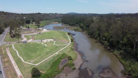 drone flying over a highway towards a green recreational park with a small brown river near the park