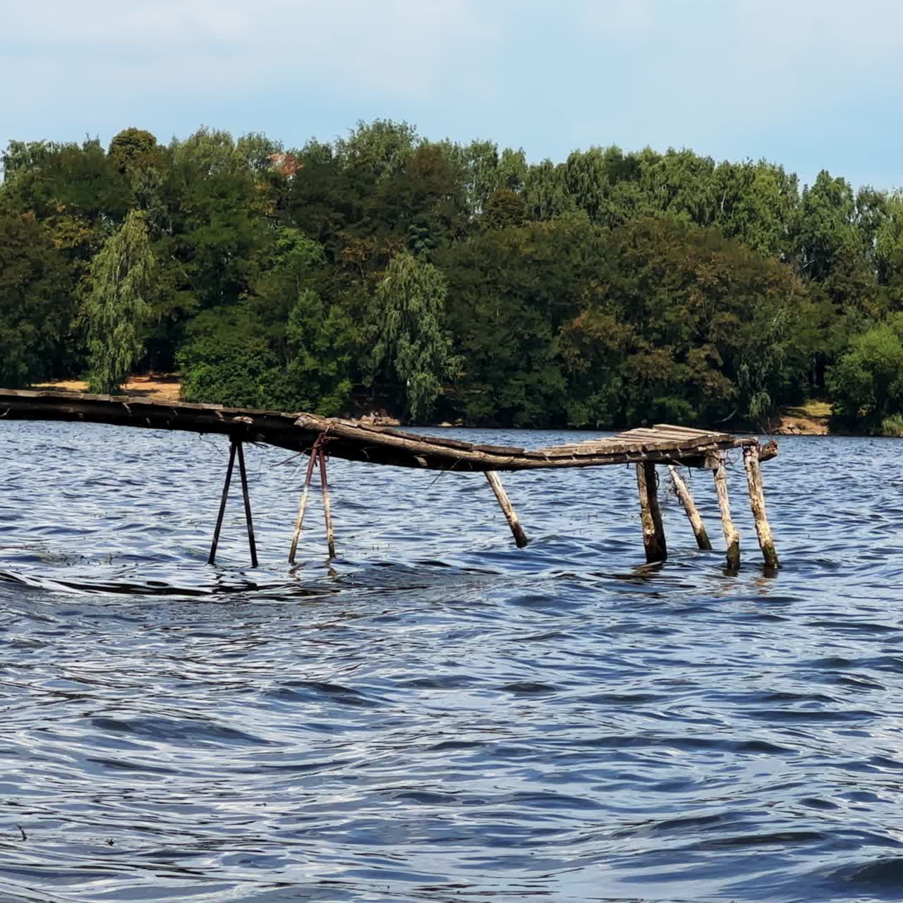 Old unsecure wooden bridge above the river. Waterfront with green trees growing at backdrop