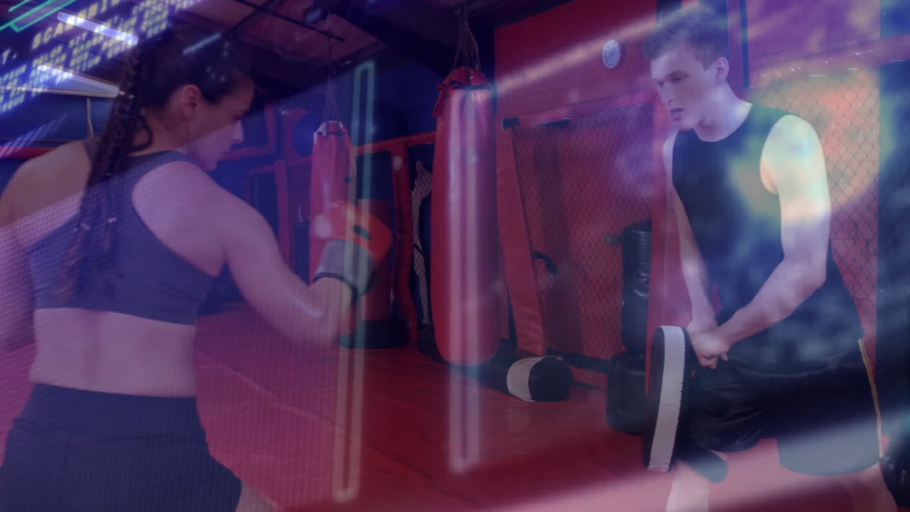 female trainer holding focus mitts coaching teenage boy in boxing gym, with neon streaks on bags