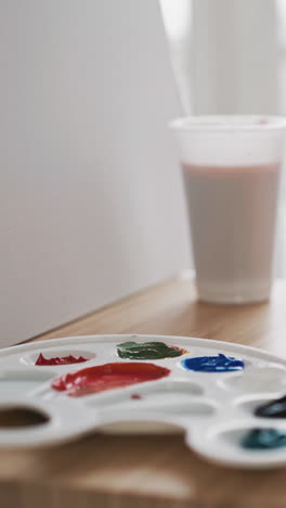 young female artist sits near palette with paints and cup of water on wooden easel with white canvas in art studio extreme close view