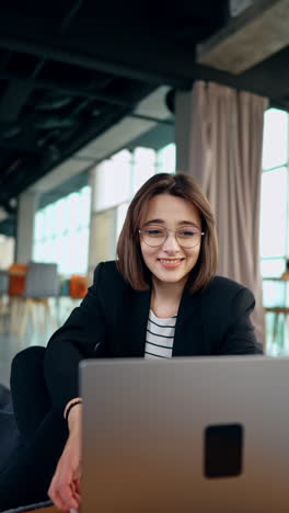 focused woman works on her computer. lady in glasses smiles to camera and puts laptop on her laps. freelance concept. vertical video.