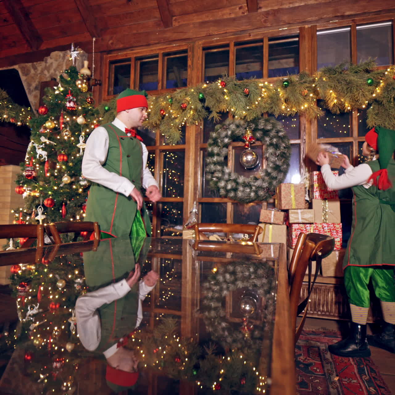 Two elves are standing in specially decorated room near the table and throwing presents to each other. Feast fireplace in the background. Christmas spirit concept close up video. Christmas eve. New Year is coming. Elves are putting presents on the table.