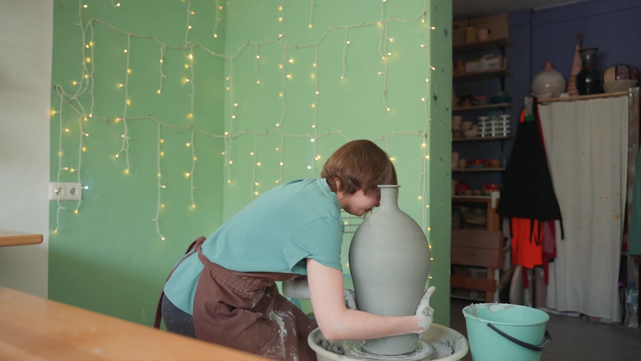 Vasaia che rifinisce il bordo di un alto vaso di argilla, scena ravvicinata in studio con parete verde e scaffali, mani che levigano e perfezionano il profilo, precisione mirata e ritmo tattile, tocchi finali