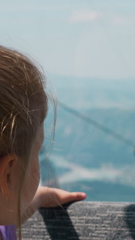 cute little girl rides modern ropeway sitting on backseat soft bench in car against distant snow capped mountains close backside view slow motion