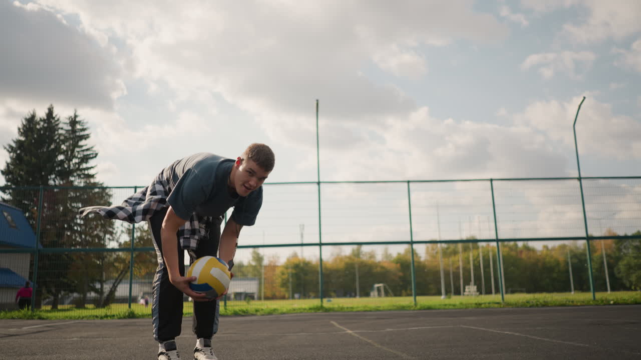 Athlete skillfully traps volleyball, putting it under arm, poses in front of camera smiling, with people walking in background on outdoor court in sports arena