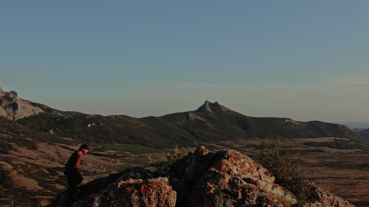 un excursionista con gorra de béisbol sube a la cima rocosa de la montaña contra el fondo del paisaje montañoso con cielo azul y puesta de sol. vista aérea. topografía por avión no tripulado con vuelo en círculo.