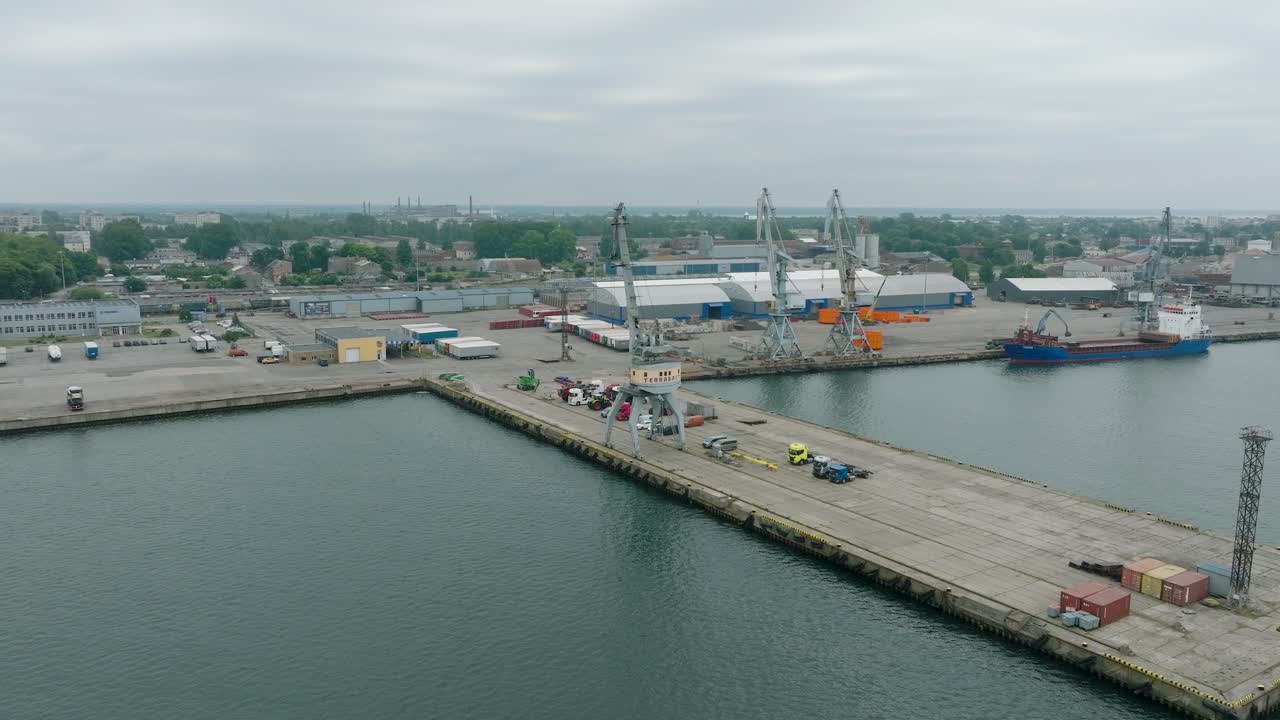 vista aérea de las grúas portuarias y los muelles de carga vacíos en el puerto de liepaja, la ciudad de liepaja en el fondo, día de verano nublado, disparo de un avión no tripulado en órbita ancha moviéndose a la derecha