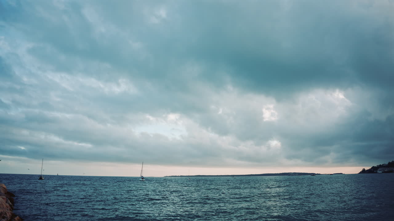 Wide shot of the sea under large clouds, with small sailboats in the distance