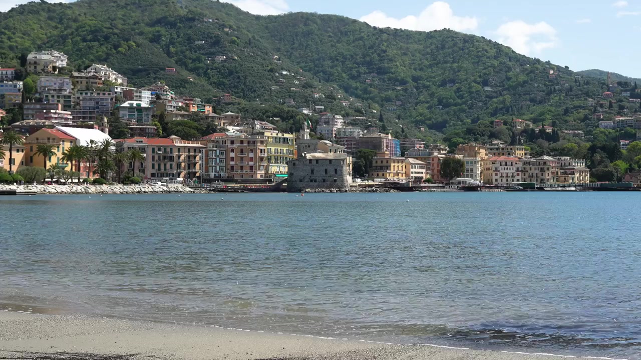 Veduta panoramica ampia di Rapallo, Italia, con edifici colorati sul lungomare, baia tranquilla, litorale sabbioso e rigogliose colline verdi sotto una luminosa luce naturale—nessuno visibile.