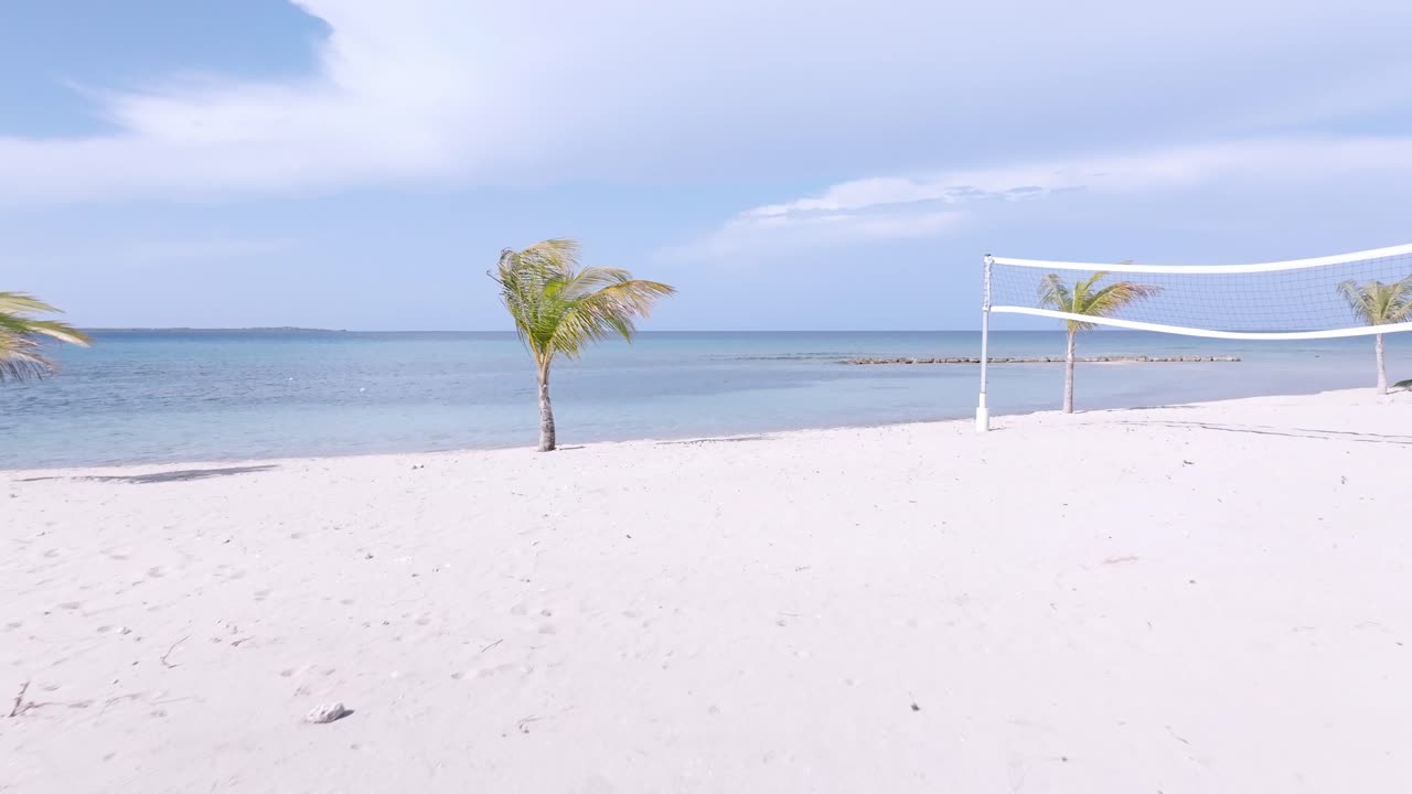 vuelo aéreo hacia adelante sobre playa de arena, campo de voleibol de playa y palmeras ondulantes frente al mar del caribe