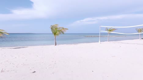 Aerial-forward-flight-over-sandy-beach,-beach-volleyball-field-and-waving-palm-trees-in-front-of-Caribbean-sea