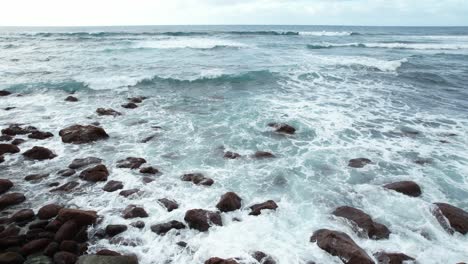 Slow-Drone-Movement-Over-A-Rocky-Shoreline-With-Foamy-Waves-Splashing