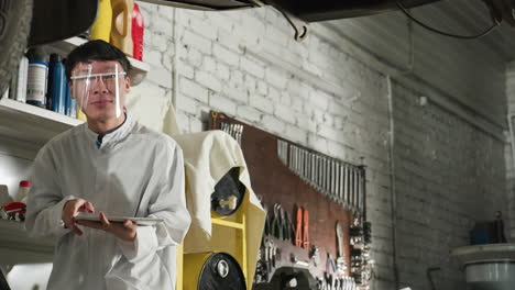 lab technician wearing face shield observes lifted car in workshop, holding tablet for technical analysis, background features mechanical equipment, industrial tools, wrenches, and repair station