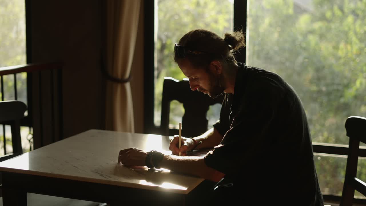 Uomo che scrive o disegna a un tavolo in una stanza illuminata dal sole