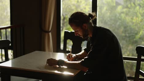 man writing or drawing at a table in a sunlit room