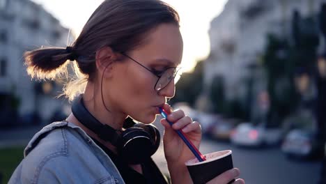 side view of a beautiful young woman with headphones on her neck drinking beverage using a straw. blonde girl in the city. son