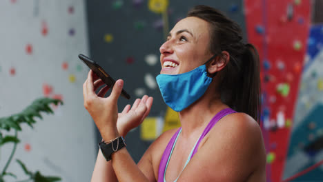 Mujer-Caucásica-Riendo-Con-Máscara-Facial-Usando-Un-Teléfono-Inteligente-En-La-Pared-De-Escalada-Interior