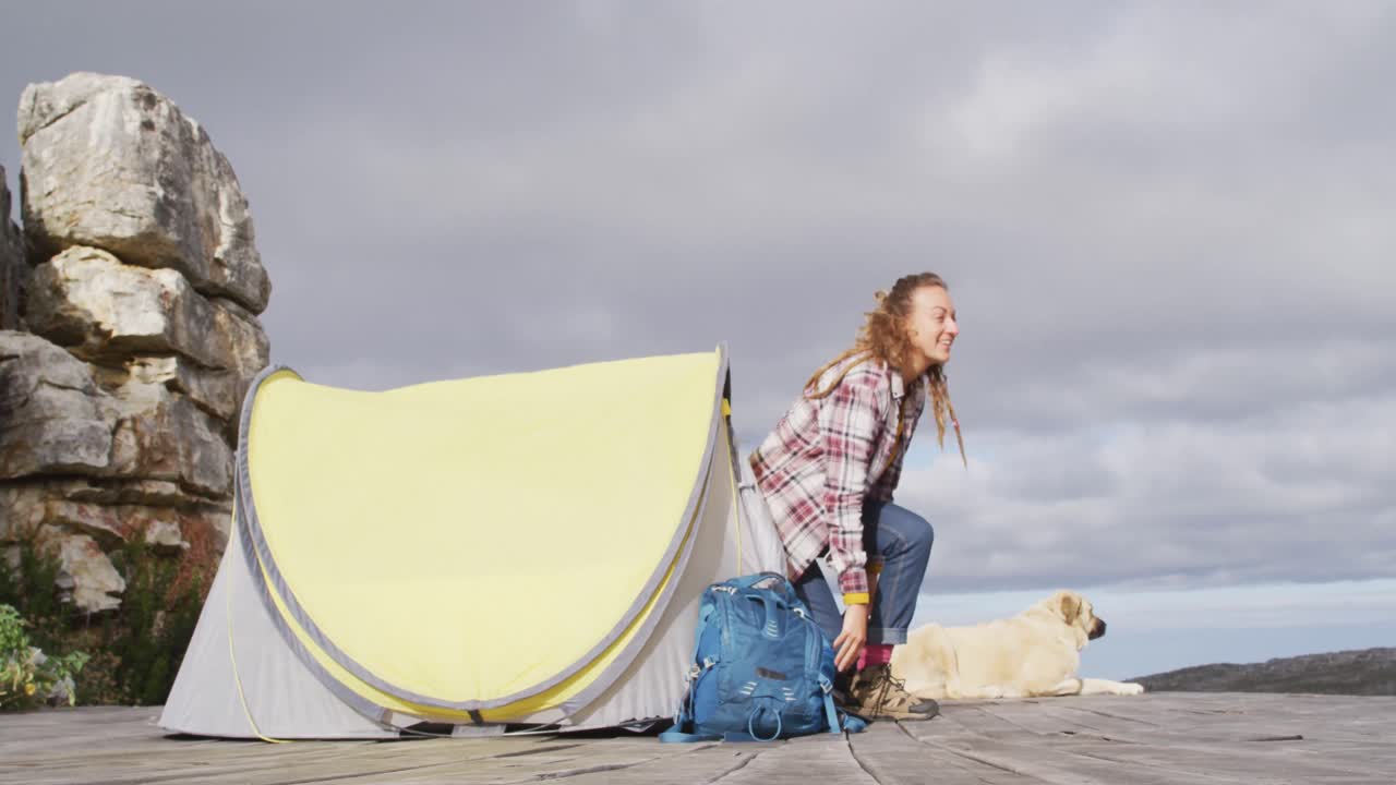 feliz mujer caucásica acampando con un perro mascota, dejando la tienda, de pie y estirándose en la ladera de la montaña