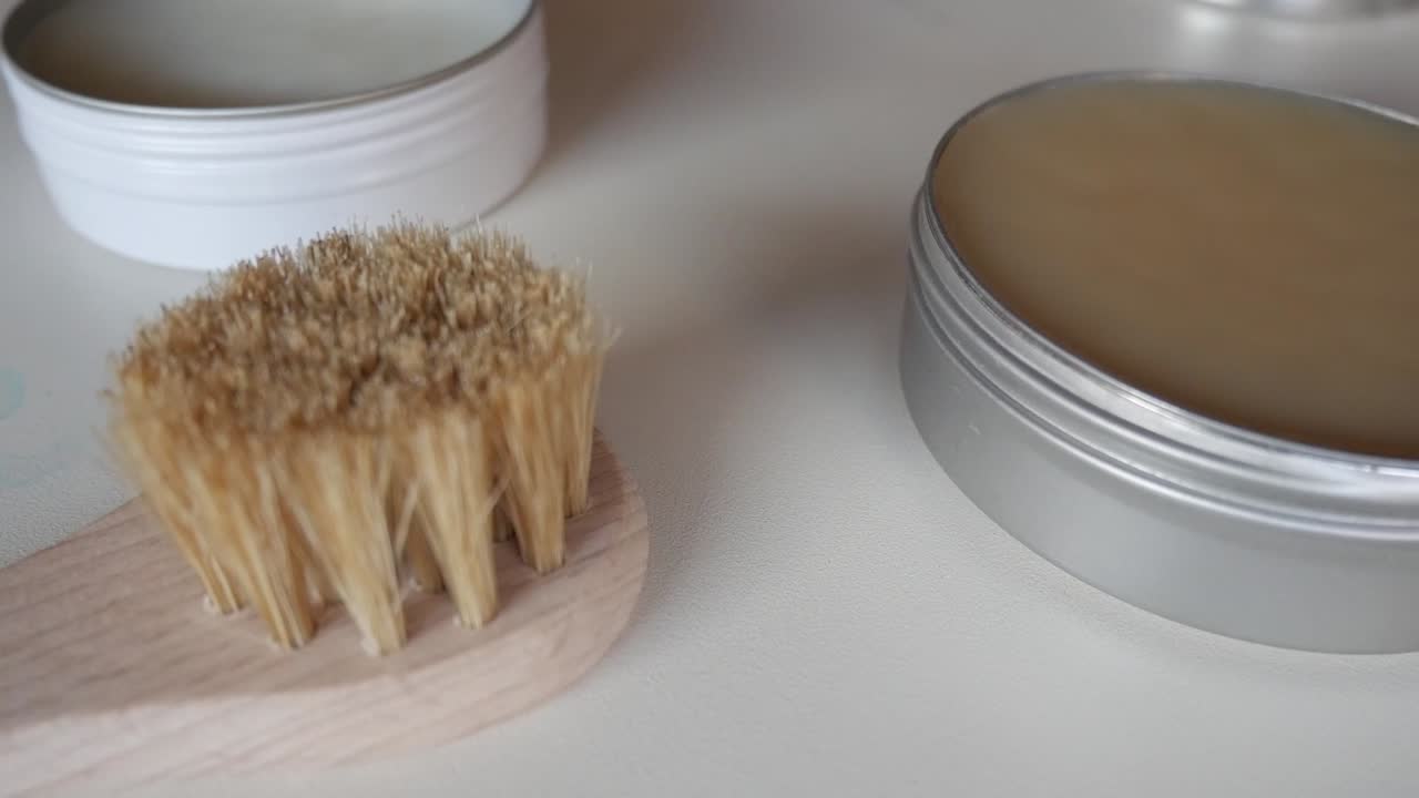Close-up of a wooden shoe polishing brush and a metal wax tin placed on a white surface under soft natural light
