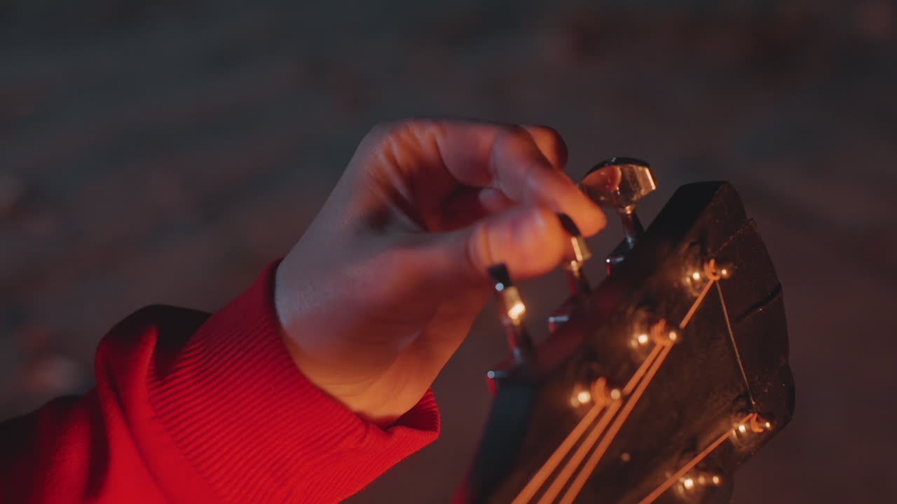 Night camping hand tuning guitar, cozy dusk scene with red sleeved camper adjusting acoustic strings, warm lantern glow, focused fingers on headstock, subtle bokeh background, relaxed outdoor mood
